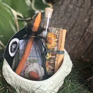 May include: A white ceramic cauldron filled with a variety of items for a spell or ritual, including a small orange candle, a glass vial, a bundle of dried herbs, and a stick of palo santo.
