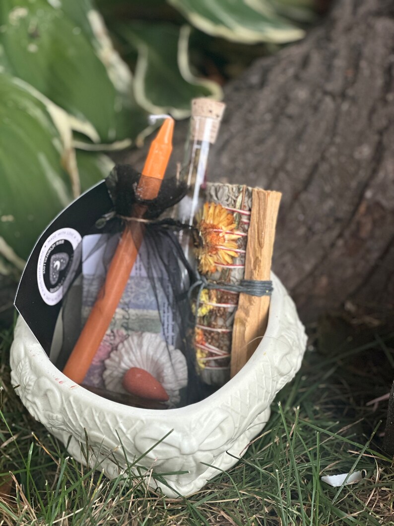May include: A white ceramic cauldron filled with a variety of items for a spell or ritual, including a small orange candle, a glass vial, a bundle of dried herbs, and a stick of palo santo.