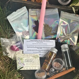May include: A collection of witchy supplies in a black cauldron, including a pink candle, small bags of herbs, a bottle of oil, a small jar of salt, a cinnamon stick, and a small vial of black powder. The text on the card reads "A blessed Reminder from your Seours Sorcieres" and "Spele".