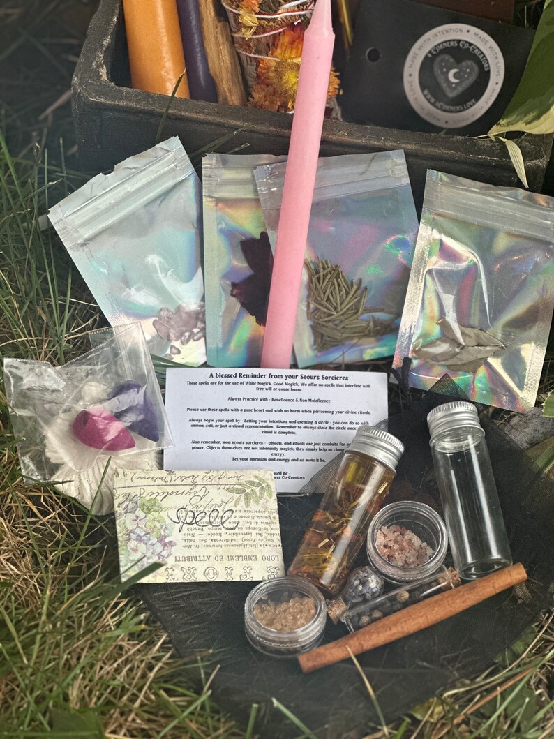 May include: A collection of witchy supplies in a black cauldron, including a pink candle, small bags of herbs, a bottle of oil, a small jar of salt, a cinnamon stick, and a small vial of black powder. The text on the card reads "A blessed Reminder from your Seours Sorcieres" and "Spele".