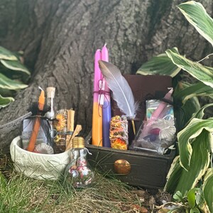 May include: A wooden box filled with various items for a witchy ritual, including candles, herbs, a feather, and a small glass bottle. The box is sitting on a bed of green grass.