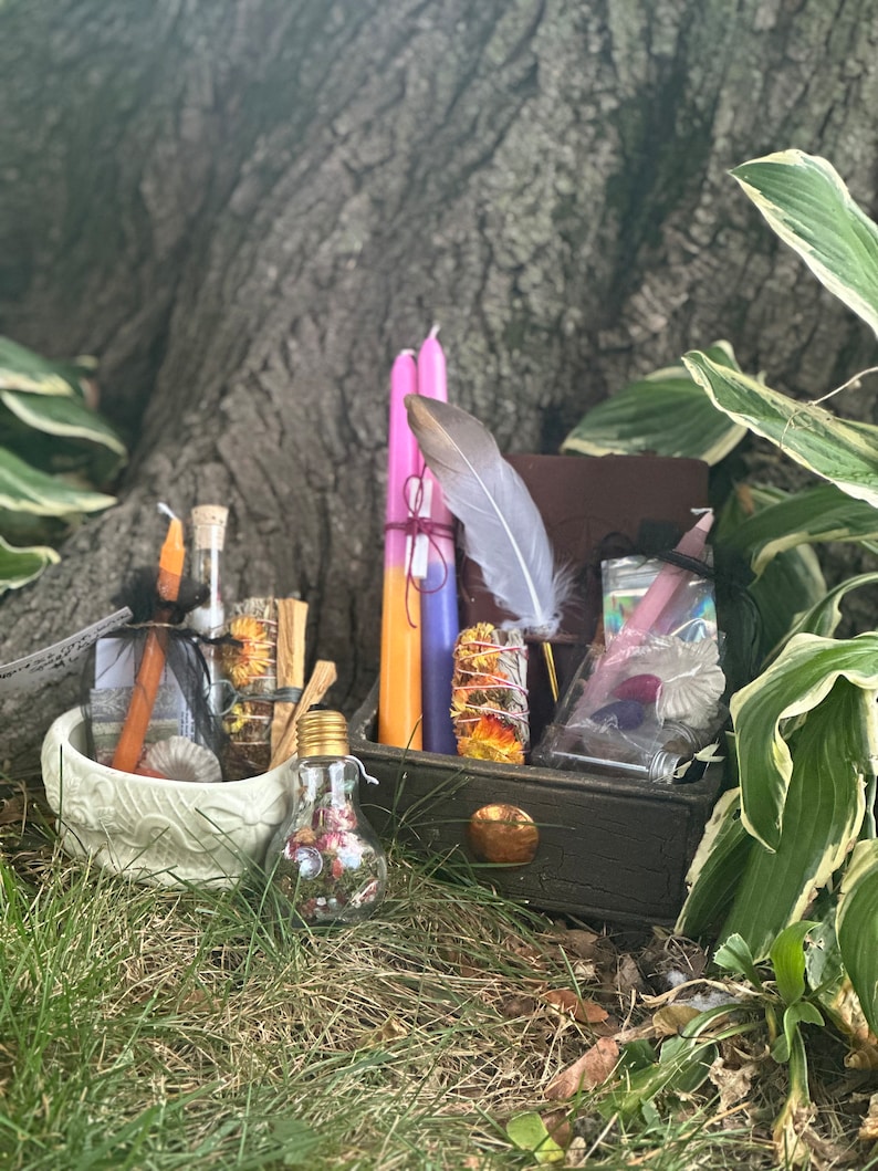 May include: A wooden box filled with various items for a witchy ritual, including candles, herbs, a feather, and a small glass bottle. The box is sitting on a bed of green grass.