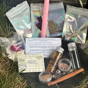 May include: A wooden box filled with various items for a spell kit, including a pink candle, small bags of herbs, a glass bottle with a cork stopper, a small jar of salt, a cinnamon stick, a small jar of brown powder, and a card with the text "Love, Embrace, & Attract".