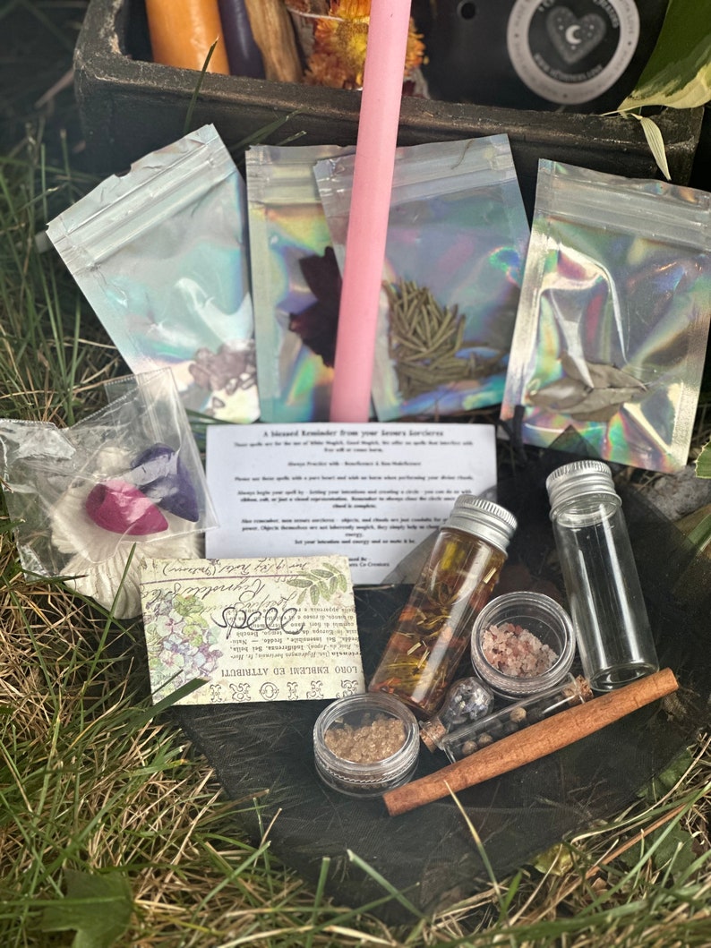 May include: A wooden box filled with various items for a spell kit, including a pink candle, small bags of herbs, a glass bottle with a cork stopper, a small jar of salt, a cinnamon stick, a small jar of brown powder, and a card with the text "Love, Embrace, & Attract".