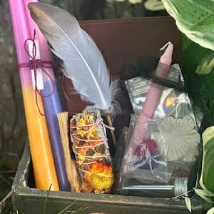 May include: A wooden box filled with various items for a spiritual ritual, including candles, a feather, a smudge stick, and a small bottle. The box is decorated with a copper-colored knob.