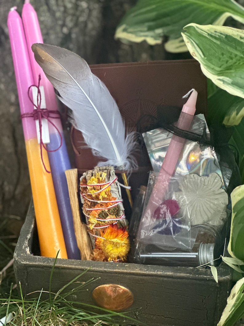 May include: A wooden box filled with various items for a spiritual ritual, including candles, a feather, a smudge stick, and a small bottle. The box is decorated with a copper-colored knob.