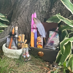 May include: A wooden box filled with various witchy items, including candles, a feather, a small bottle, and a bowl. The box is sitting on a bed of green grass.