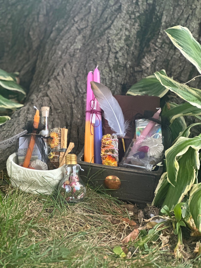 May include: A wooden box filled with various witchy items, including candles, a feather, a small bottle, and a bowl. The box is sitting on a bed of green grass.