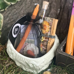 May include: A white ceramic bowl with a floral design, filled with a variety of items, including a wooden stick, a glass vial with dried flowers, a bundle of dried herbs, and a small orange candle. The bowl is sitting on a bed of green grass.