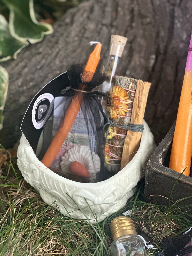 May include: A white ceramic bowl with a floral design, filled with a variety of items, including a wooden stick, a glass vial with dried flowers, a bundle of dried herbs, and a small orange candle. The bowl is sitting on a bed of green grass.