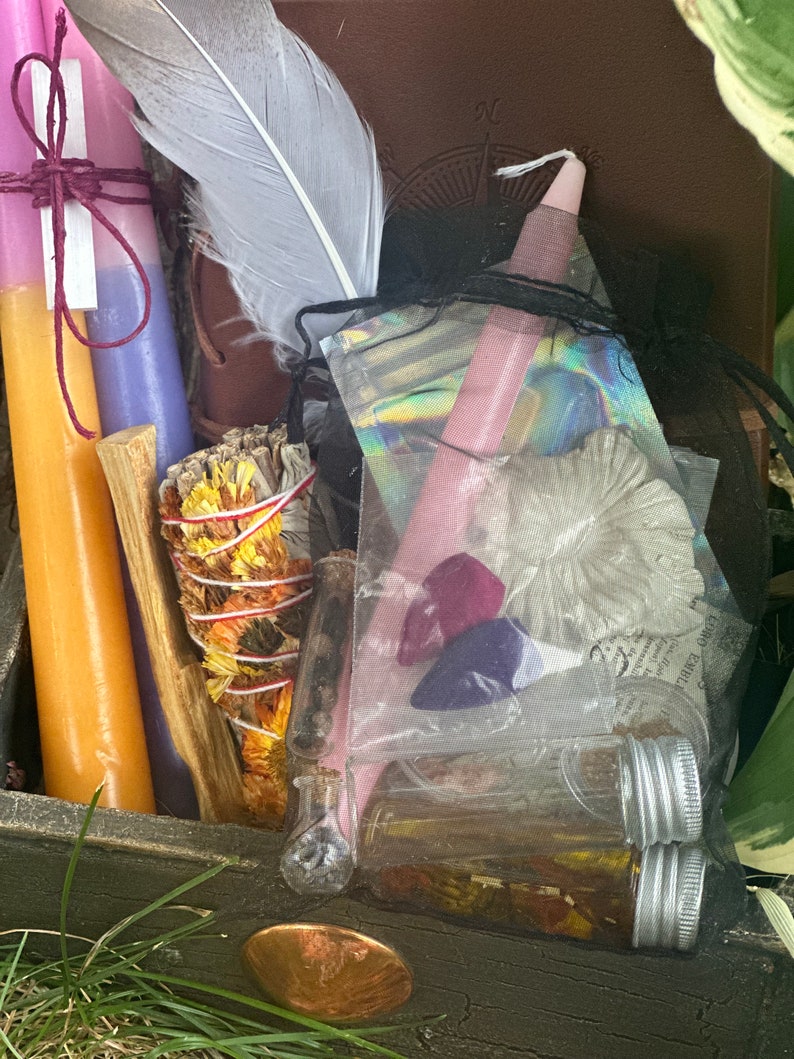 May include: A wooden box filled with various items for a spiritual ritual, including candles, sage, crystals, and a feather. The box is decorated with a copper coin.