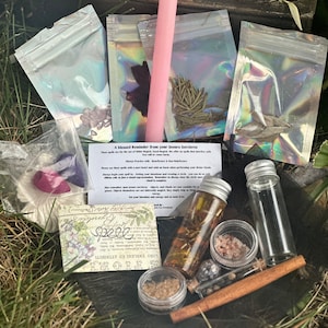 May include: A collection of witchy supplies in a wooden box, including a pink candle, small bags of herbs, a glass bottle of oil, a small jar of salt, a cinnamon stick, and a small glass vial. The box is sitting on a bed of green grass. The text on the card reads "A blessed Reminder from your Seours Sorcieres".