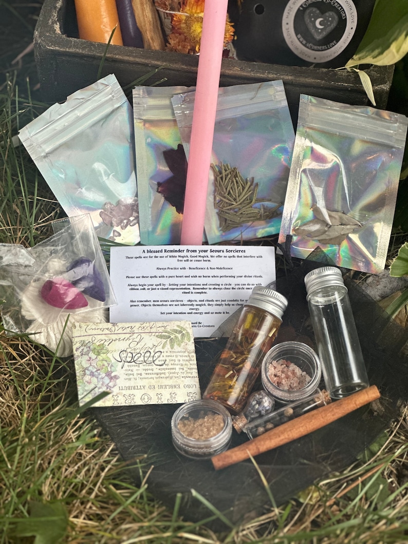 May include: A collection of witchy supplies in a wooden box, including a pink candle, small bags of herbs, a glass bottle of oil, a small jar of salt, a cinnamon stick, and a small glass vial. The box is sitting on a bed of green grass. The text on the card reads "A blessed Reminder from your Seours Sorcieres".