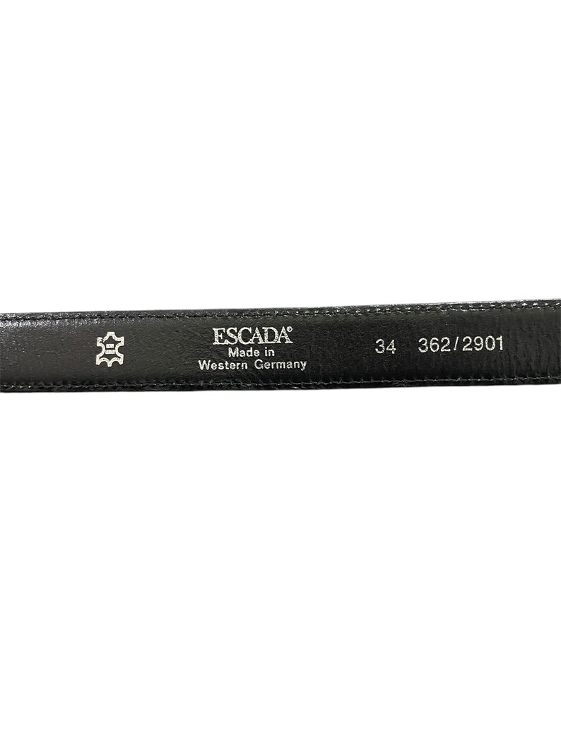 Op de afbeelding: Zwarte leren riem met een zilveren gesp en de tekst "ESCADA Made in Western Germany 34 362/2901" gedrukt op de achterkant.