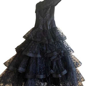 Puede incluir: Un vestido de tul negro con escote de un hombro y falda de capas. La falda está adornada con lunares negros.