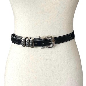Op de afbeelding: Een zwarte leren riem met een zilveren gesp en drie zilveren decoratieve stukken. De riem is op een wit mannequin.
