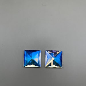 May include: Two square-cut, iridescent blue crystals. The crystals have a multi-faceted design that reflects light, creating a spectrum of colors. The crystals are set against a gray background.