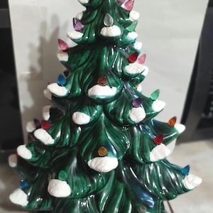 May include: A green ceramic Christmas tree with a glossy finish, featuring textured white snow accents on the branches and numerous small, multi-colored translucent plastic light bulbs inserted throughout the structure on a round base.