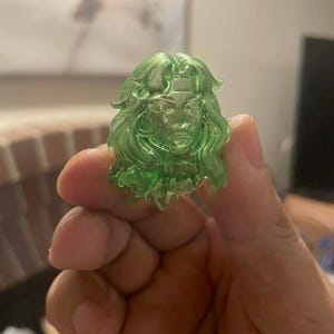 May include: A translucent green figurine of a woman's head with long, wavy hair. The figure has detailed facial features and is held in a person's hand. The material appears to be a type of resin or plastic.
