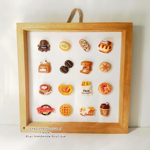 Puede incluir: Un marco de madera con fondo blanco muestra una colección de artículos de comida en miniatura, incluyendo pasteles, café y un pequeño letrero de tienda. Los artículos están hechos de arcilla polimérica y están dispuestos en un patrón de cuadrícula. El marco está decorado con un asa de cuerda marrón.