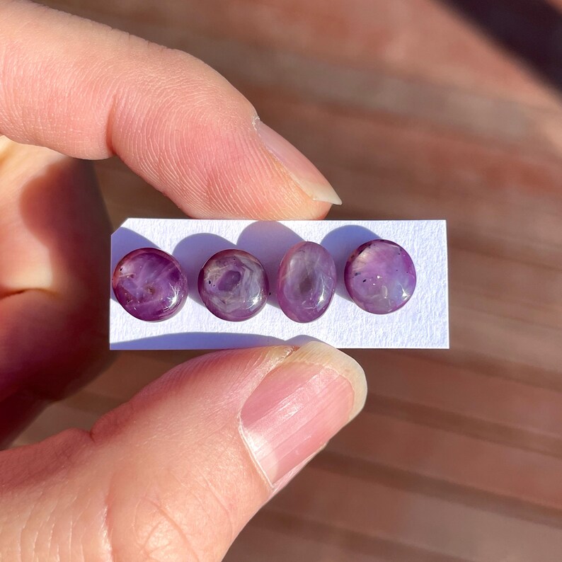 May include: Four polished, oval-shaped gemstones with a deep purple hue, displayed on a white card. The stones are held between fingers, showcasing their translucent quality and unique internal patterns. The gemstones are approximately 0.5 inches in diameter.