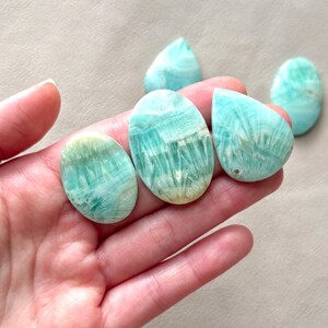 May include: A hand holds several polished gemstones in various shapes, including oval and teardrop. The stones display a mix of light blue and white colors with a natural, textured pattern. The gemstones are likely used for jewelry or decorative purposes.