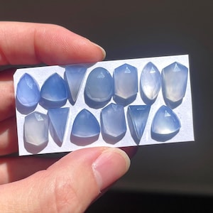 May include: A collection of 14 blue chalcedony cabochons in various shapes and sizes, displayed on a white card.
