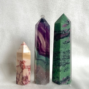 May include: Three gemstone obelisks of different sizes and colours. The smallest is cream and red, the middle is purple and white, and the largest is green with red inclusions. All have a polished, faceted design and stand on a white surface.