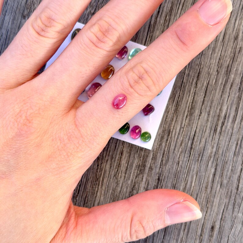 May include: A collection of small, oval-shaped, multi-colored cabochon gemstones arranged on a white display card. The stones feature various translucent hues including pink, green, yellow, and red, each polished to a smooth, rounded finish.