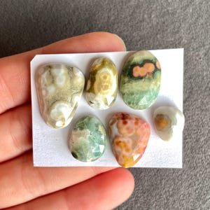 May include: Six polished gemstones in assorted shapes and colours. The stones display patterns of beige, green, orange, and white. Some stones have faceted edges, and all are presented on a white card.