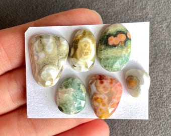 May include: A collection of six polished gemstones in various shapes and colors. The stones feature patterns of beige, green, orange, and white. Some stones have faceted edges, and all are displayed on a white card.