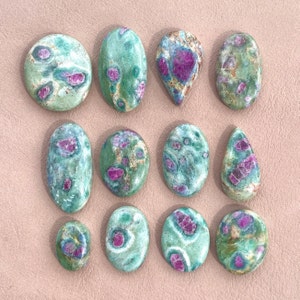 May include: Twelve green and pink ruby zoisite cabochons, each with a unique pattern of pink and green. The stones are smooth and polished, and they are arranged in a grid pattern.