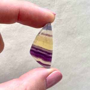 May include: A triangular, yellow, purple, and white fluorite cabochon gemstone. The gemstone is translucent and has a layered pattern.