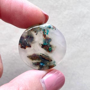 May include: A round, white agate cabochon with inclusions of copper and turquoise. The cabochon is approximately 25mm in diameter.