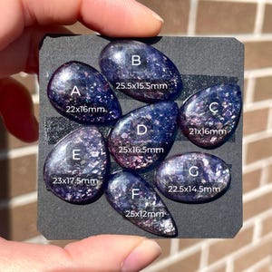 May include: Seven polished, oval and teardrop-shaped gemstones with a dark blue and purple speckled appearance. Each stone is labeled with a letter and size in millimeters, ranging from 21x16mm to 25.5x16.5mm. The gemstones are displayed on a black surface.