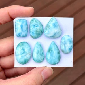 May include: Eight polished larimar gemstones in various shapes, including oval, teardrop, and rectangular. The stones display a range of blue and white hues, with unique patterns. The gemstones are displayed on a white card.