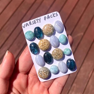 May include: A selection of polished gemstones in shades of blue, green, and brown, presented on a white card. The card is labelled "VARIETY PACKS" in handwritten text. The gemstones are oval and round.