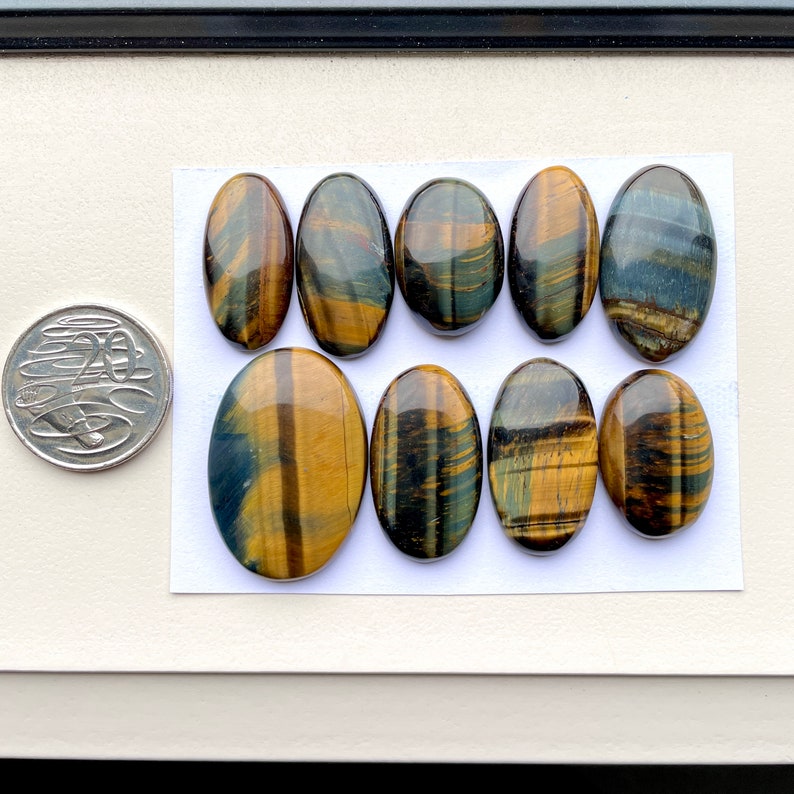 May include: Ten oval-shaped tiger's eye gemstones with a mix of golden brown and blue stripes. The stones are arranged in two rows on a white surface.