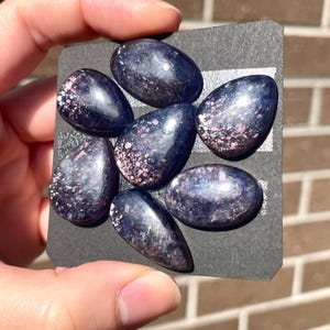May include: Seven polished, oval-shaped gemstones in shades of deep blue and purple, with sparkling pink and white inclusions. The stones are arranged on a black card, showcasing their varied shapes and textures. The gemstones are held in a hand.