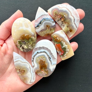 May include: A collection of polished agate stones in various shapes, including oval, teardrop, and rectangular. The stones feature intricate patterns of white, brown, green, and red, creating a natural, earthy aesthetic. The stones are held in a hand.