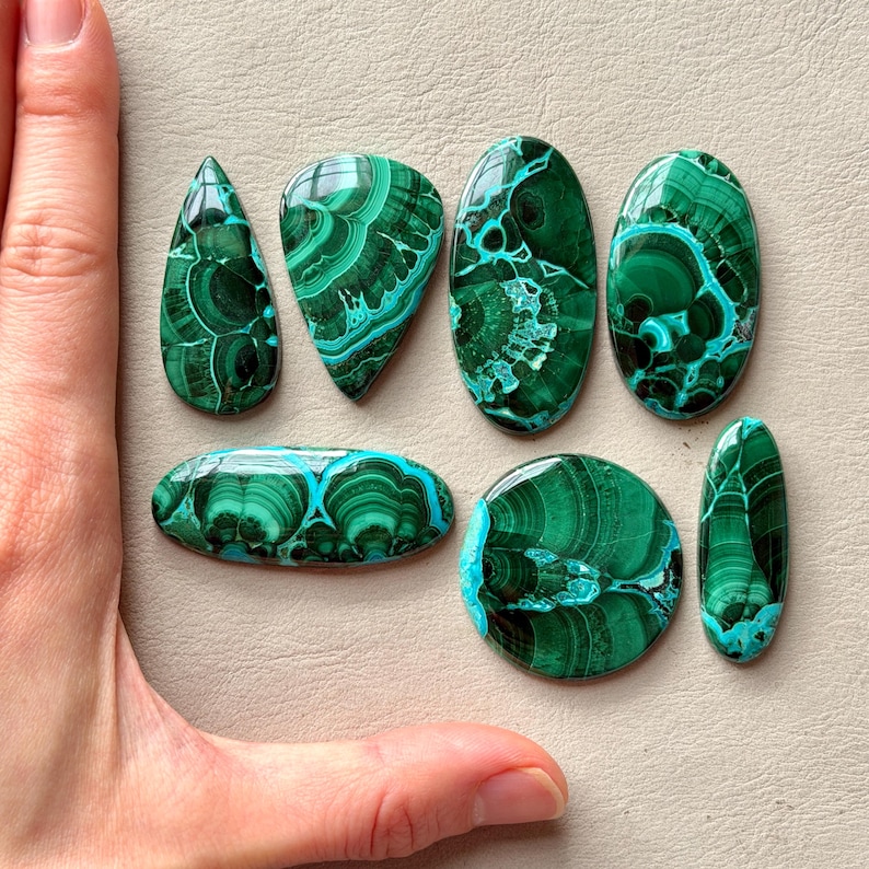 Pu&ograve; includere: Una collezione di sei pietre preziose in malachite lucida di varie forme, tra cui goccia, ovale e circolare. Ogni pietra presenta motivi a bande verde scuro e verde acqua con vortici, anelli concentrici e texture organiche stratificate.