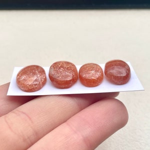 Peut inclure: Quatre cabochons ovales en pierre de soleil, chacun avec un motif d'inclusions diff&eacute;rent, pos&eacute;s sur un fond blanc.