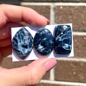 Könnte beinhalten: Drei blaue Tigeraugen-Cabochons, ein ovaler, ein rechteckiger und ein tropfenförmiger. Die Steine haben einen schimmernden, irisierenden Effekt.