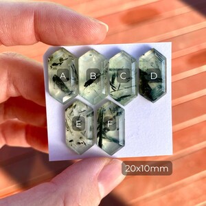 K&ouml;nnte beinhalten: Sechs hexagonale Prehnit-Edelsteine mit dunkelgr&uuml;nen Einschl&uuml;ssen werden gezeigt. Jeder Stein ist mit einem Buchstaben von A bis F gekennzeichnet. Die Abmessungen betragen 20x10mm. Die Edelsteine werden vor einem wei&szlig;en Hintergrund gehalten.