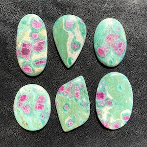 May include: Six polished gemstones in various shapes, featuring a vibrant mix of colours. The stones showcase a teal base with striking magenta and white patterns. The collection includes oval, teardrop, and irregular shapes, ideal for jewellery making.