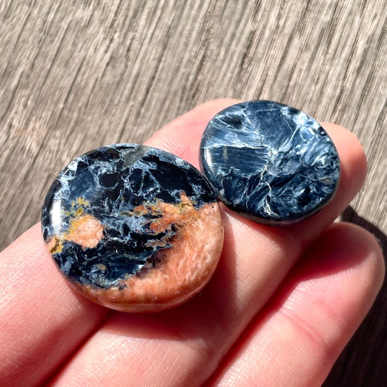 May include: Two round, polished stones with a blue and black tiger's eye pattern. One stone has a pink and orange color variation.