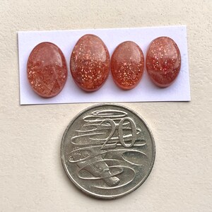 Peut inclure: Quatre cabochons ovales en pierre de soleil de couleur rouge-orang&eacute; avec une finition scintillante. Les pierres sont dispos&eacute;es en rang&eacute;e sur un fond blanc. Une pi&egrave;ce australienne de 20 cents est pr&eacute;sent&eacute;e pour comparaison de taille.