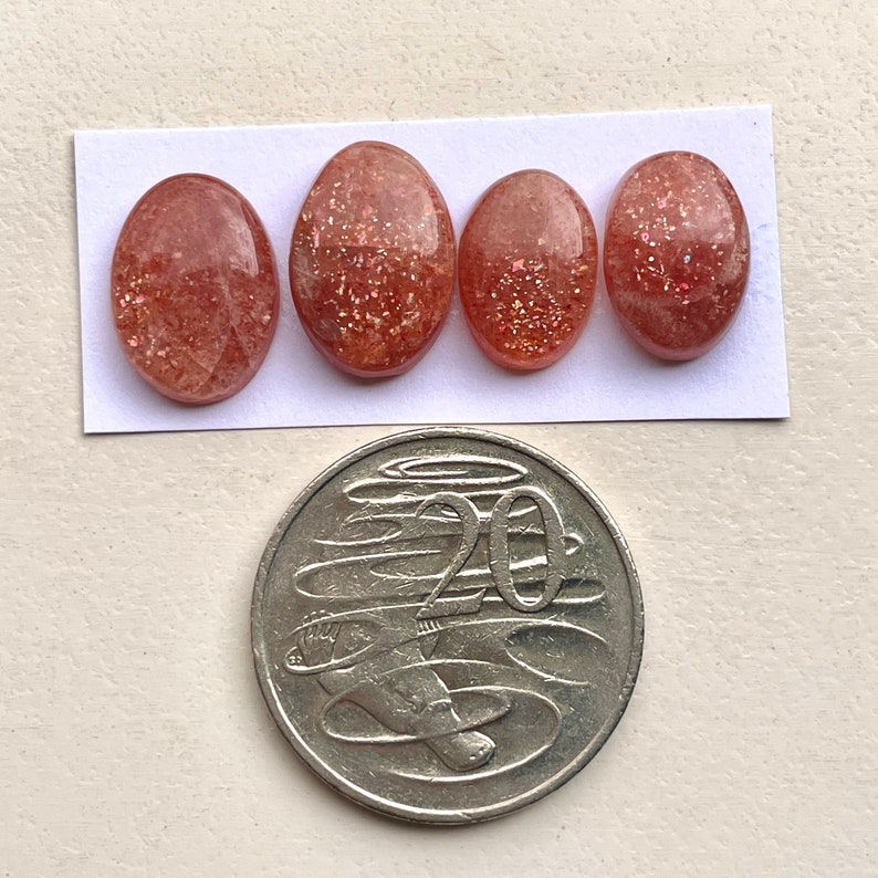 Peut inclure: Quatre cabochons ovales en pierre de soleil de couleur rouge-orang&eacute; avec une finition scintillante. Les pierres sont dispos&eacute;es en rang&eacute;e sur un fond blanc. Une pi&egrave;ce australienne de 20 cents est pr&eacute;sent&eacute;e pour comparaison de taille.
