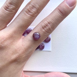 Pode incluir: Uma cole&ccedil;&atilde;o de pedras preciosas polidas, redondas, com corte caboch&atilde;o, em tons de roxo e rosa. As pedras s&atilde;o exibidas em uma superf&iacute;cie branca, mostrando suas superf&iacute;cies lisas e reflexivas. As pedras t&ecirc;m aproximadamente 0,6 cm de di&acirc;metro.