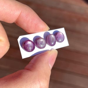 May include: Four oval-shaped, polished gemstones in varying shades of purple and pink are displayed on a white card. The gems have a smooth, reflective surface, and are held between fingers. The background is blurred.